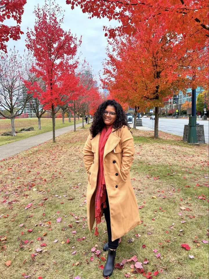Sanchita smiling in a camel coat and red scarf, walking beneath red and orange autumn trees
