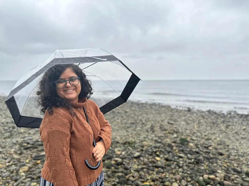 Sanchita smiling under a clear umbrella on a pebble beach on a grey, rainy day