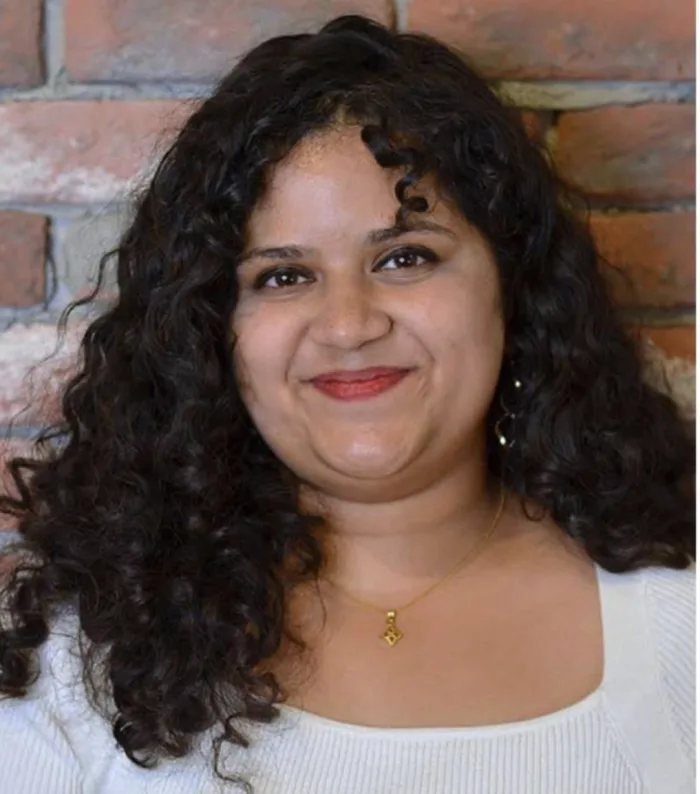 Sanchita smiling, with long curly dark hair, in front of a brick wall