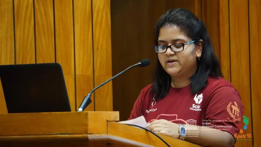 Sanchita speaking at a podium in a Scio Foundation t-shirt